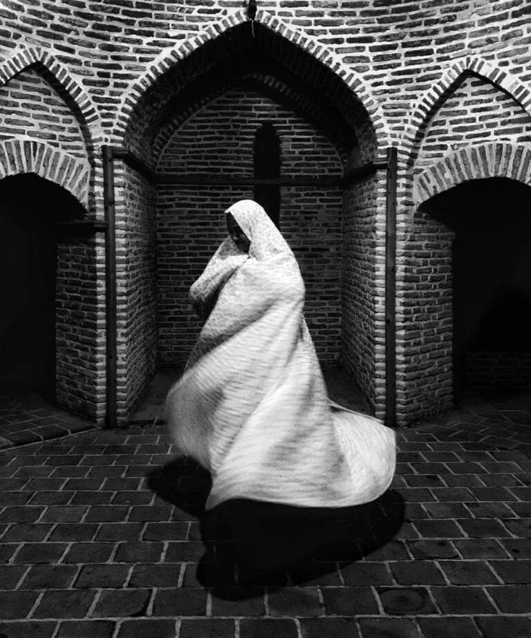 Donna in velo di stoffa, scena in bianco e nero, architettura in mattoni antica, atmosfera sacra e mistica.