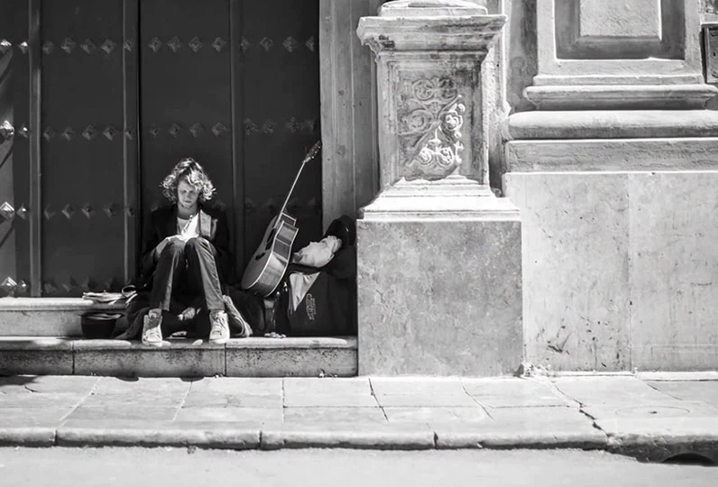 Due persone senza fissa dimora sedute su un marciapiede con una chitarra.