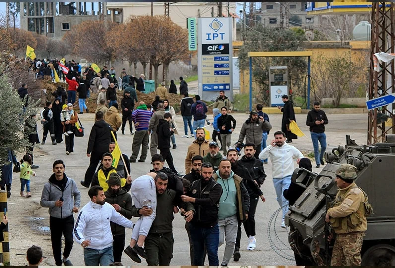Gruppo di rifugiati e migranti che camminano lungo una strada con forze militari presenti.